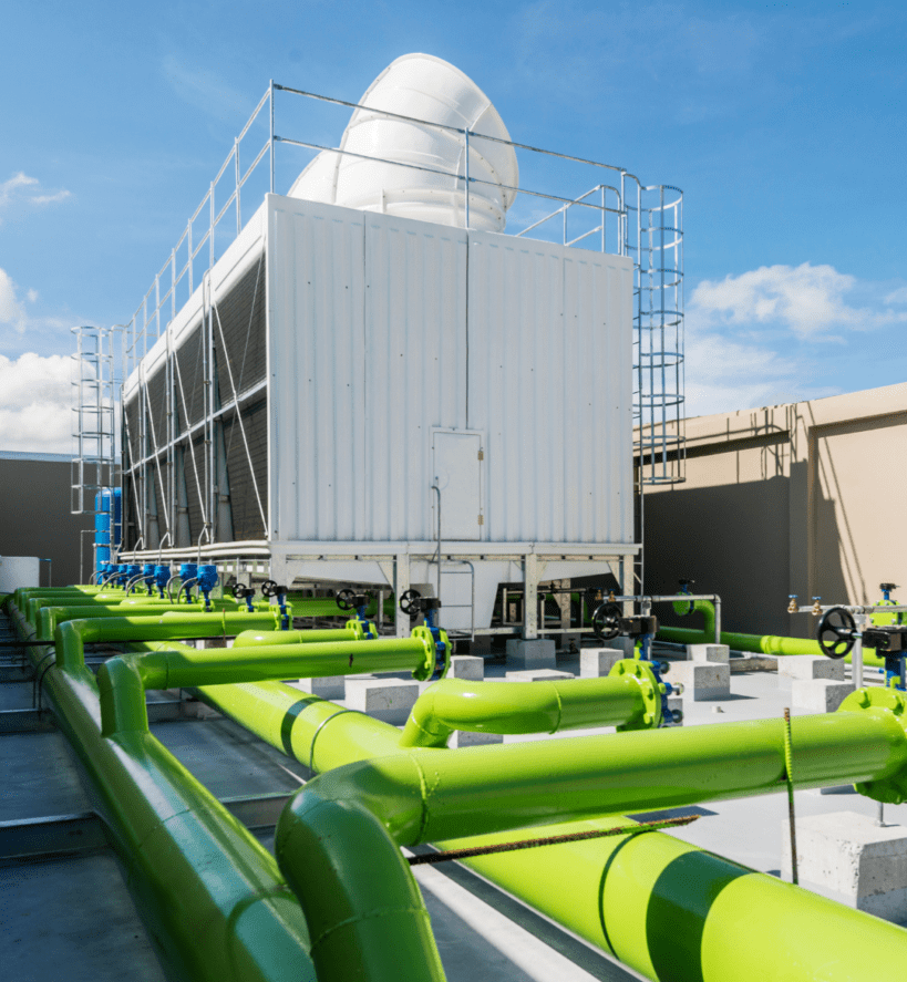 Cooling-Tower-819x1024 Rooftop cooling tower on a commercial building with bright green water piping, valves, and blue pumps under a clear blue sky, with the Depue Mechanical logo in the corner.
